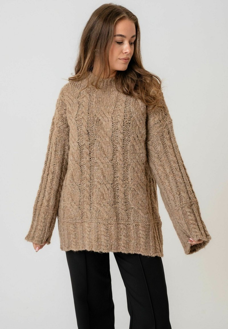 Prevelik, teksturirani pleteni pulover u svijetlosmeđoj boji s uzorcima pletenice. Ima duge rukave i široki rebrasti rub. Uparen s crnim hlačama.