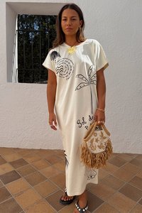 Abito maxi color crema chiaro con grafiche di palme nere e testo. Presenta una scollatura rotonda e maniche corte. Gli accessori includono una borsa intrecciata.