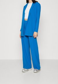 Blå blazer och matchande vida byxor i slätt tyg, kombinerat med en vit kort topp och silvriga sneakers.