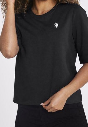 Vrouw draagt een zwart T-shirt met korte mouwen en een klein wit geborduurd logo op de linkerborst, houdt de zoom voorzichtig vast.