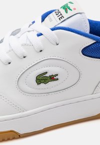 Bílá teniska s modrými detaily, bílými tkaničkami, gumovou podrážkou a zeleným krokodýlem Lacoste na boku a štítku na jazyku.