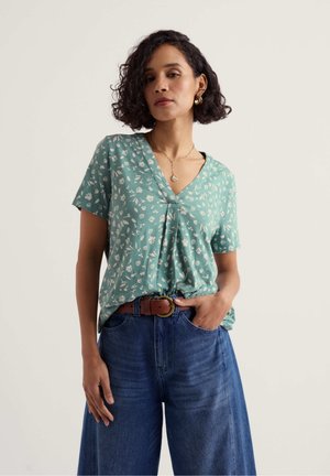 Woman with curly hair wearing green floral V-neck shirt, blue high-waist wide-leg jeans, brown belt, gold earrings, and pendant necklace.