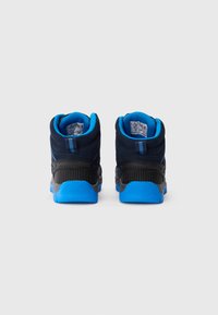 Pár navy a černých turistických bot s jasně modrými podrážkami a interiérem, z pohledu ze zad na světlém jednolitém pozadí.