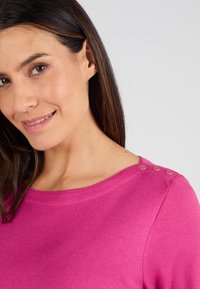 Top rosa con un escote amplio, que presenta tres botones decorativos en el hombro. Textura suave y ajuste casual, adecuado para el uso diario.