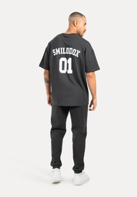 Schwarzes oversized T-Shirt mit "SMILODOX 01" in Weiß auf der Rückseite, kombiniert mit dunklen Jogginghosen und weißen Sneakers, vor einem weißen Hintergrund stehend.