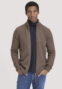 Brauner Reißverschluss-Pullover mit hohem Kragen, gestricktem Muster, gerippten Bündchen und Saum. Über einem dunklen Rollkragenpullover getragen und mit blauen Jeans kombiniert.