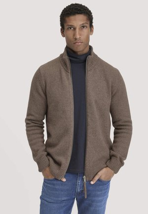 Brown zip-up sweater with a high collar, knitted texture, ribbed cuffs, and hem. Worn over a dark turtleneck and paired with blue jeans.