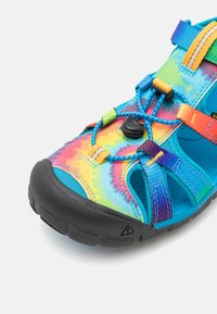 Farebná sandála s technikou tie-dye, vybavená gumovou podrážkou, nastaviteľnými popruhmi a regulátorom na prispôsobenie. Vrchná časť má živé odtiene modrej, ružovej a žltej.