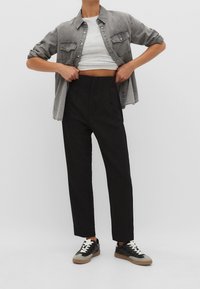 Grijze denimshirt gelaagd over een witte cropped top, gecombineerd met hoge zwarte broek en zwarte sneakers met witte veters en bruine accenten.