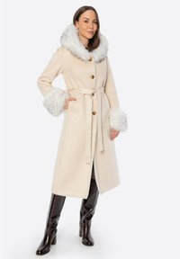Beżowy płaszcz długości do kolan z dużym białym futerkiem z sztucznego włosia na kołnierzu i mankietach, z paskiem w talii, guzikami i miękką teksturą. Brązowe botki na wysokim obcasie.