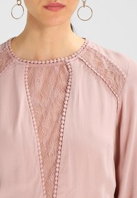 Ljusrosa blus med spetsdetaljer längs halsringningen, texturerat tyg och dekorativa pom-pom accenter runt kragen och axlarna.