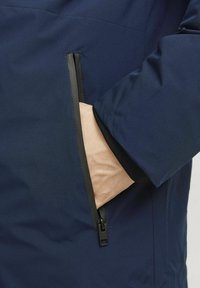 Veste légère bleu marine avec une poche zippée, des accents noirs et une texture douce, mettant en avant un design épuré.