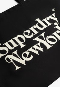 Czarna torba na ramię z płótna z wyraźnym białym napisem "Superdry New York". Wytrzymały materiał, długie uchwyty i prostokątny kształt.
