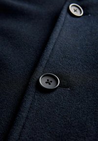Temno modri plašč iz tkanine v bližnjem načrtu, z črnim gumbom s štirimi luknjami, gladko teksturo in vidnimi šivi vzdolž prednjega dela.