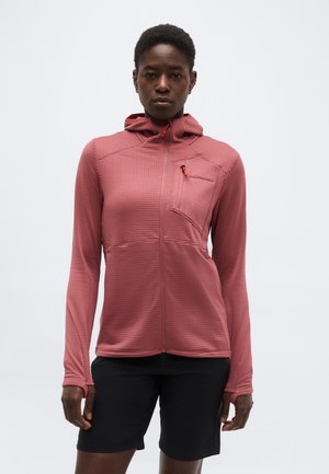 Asmuo dėvintis dulkinės rožės spalvos su užtrauktuku gobtuvinę striukę su krūtinės kišene ir juodus kelio ilgio šortus, stovintis priešais paprastą foną.