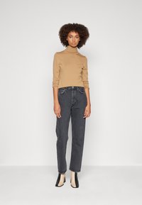 Beige garbó pulóver, fekete magas derekú farmernadrág és kéttónusú bokacsizma. A modell egyenesen áll egy sima fehér háttér előtt.