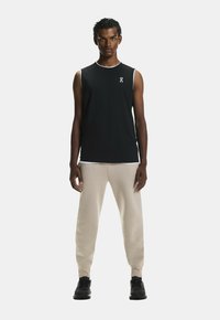 Schwarzes ärmelloses Sporttop mit weißem Saum und Logo, kombiniert mit beigen, schmal zulaufenden Jogginghosen und schwarzen Sportschuhen.