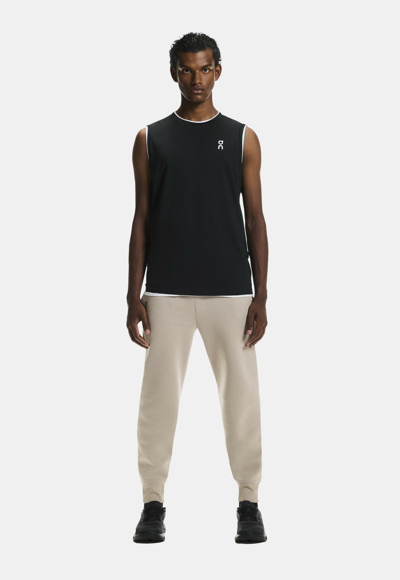 Schwarzes ärmelloses Sporttop mit weißem Saum und Logo, kombiniert mit beigen, schmal zulaufenden Jogginghosen und schwarzen Sportschuhen.