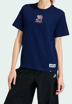 Persona que lleva una camiseta azul marino con bordado de temática de tenis y pantalones negros Adidas con rayas blancas.