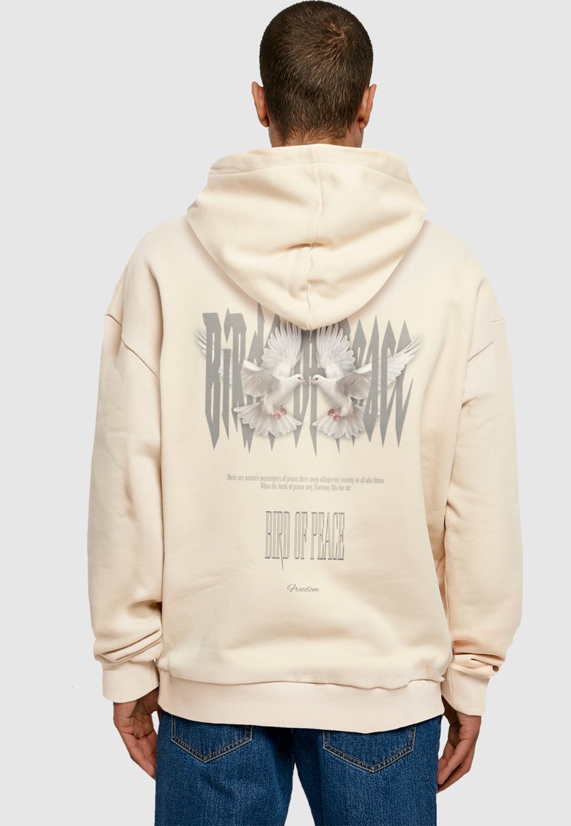 Beige oversized hoodie met een grafisch ontwerp van duiven en vetgedrukte tekst "VOGEL VAN VREDE." Zachte stof met ribgebreide boorden en onderrand.