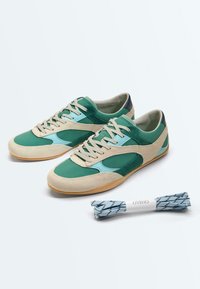 Paire de baskets en daim vert et beige avec semelles en caoutchouc, accompagnée d'un lot de lacets bleu et blanc à motif portant l'étiquette "OYSHO".