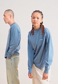 Long-sleeve blue cotton shirts worn by two people; one has a small "obey" logo on the chest. Light-colored pants and braided hair details visible.