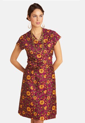 Frau trägt ein lila geblümtes Kleid mit gelben Blumen, kombiniert mit geschichteten goldenen Halsketten und goldenen Creolen, Hände hinter dem Rücken.