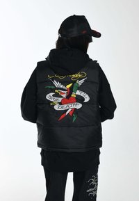 Schwarze Steppweste mit Kapuze, die ein bunt gesticktes Grafikdesign auf dem Rücken aufweist, mit einem Herz, einem Vogel und dem Text "TRUE TIL DEATH".