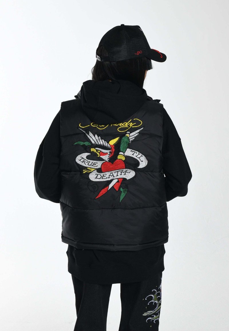 Schwarze Steppweste mit Kapuze, die ein bunt gesticktes Grafikdesign auf dem Rücken aufweist, mit einem Herz, einem Vogel und dem Text "TRUE TIL DEATH".