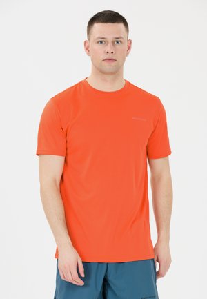 Ung mand iført en orange kortærmet sportstrøje og blå shorts, stående foran en ensfarvet hvid baggrund, med fronten vendt mod kameraet.