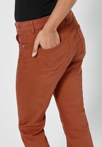 Pantalon marron, coupe slim, en tissu doux avec un détail de poche arrière et un design de jambe légèrement effilé. Présente des surpiqûres visibles et une boucle de ceinture.