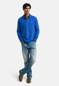 Blauw poloshirt met lange mouwen, ontspannen pasvorm, gecombineerd met licht gewassen spijkerbroek en bruine enkellaarzen. Bevat een kraag en logo-detail.