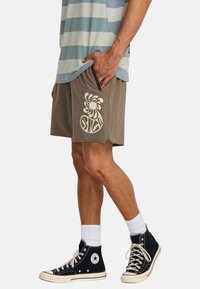 Pantaloni corti marroni con una grafica a spirale bianca, realizzati in materiale morbido. Abbinati a sneaker alte nere e una maglietta a righe blu-verdi.