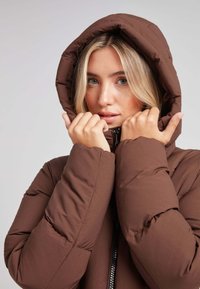 Chaqueta acolchada marrón con capucha, que cuenta con cierre de cremallera y una textura gruesa y acolchada. El modelo sostiene la capucha cerca de la cara.