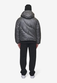 Grijze gewatteerde jas met een golfpatroon, capuchon en elastische manchet, gecombineerd met zwarte broek en witte sneakers, gezien vanuit de achterkant.