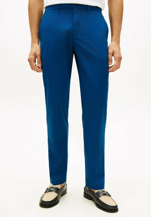 Mężczyzna noszący dopasowane niebieskie spodnie garniturowe z bocznymi kieszeniami oraz czarno-beżowe mokasyny, stojący na białym tle.