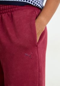Bordeauxfarvede joggerbukser lavet af blødt stof, med elastisk talje, sidelommer og et lille lilla Puma-logo broderet på låret.