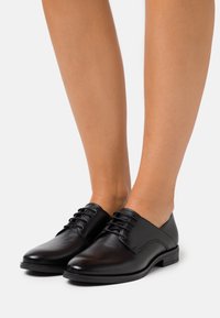Scarpe in pelle nera con lacci, dalla finitura liscia, punta arrotondata, tacco basso e dettagli cuciti sui lati.