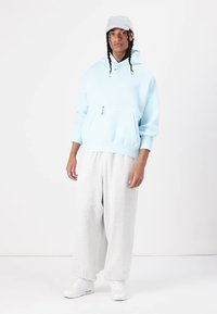 Ανοιχτό μπλε φούτερ με φερμουάρ στην μπροστινή τσέπη, συνδυασμένο με γκρίζα φαρδιά φόρμα. Το μοντέλο φοράει γκρίζο καπέλο και λευκά αθλητικά παπούτσια.