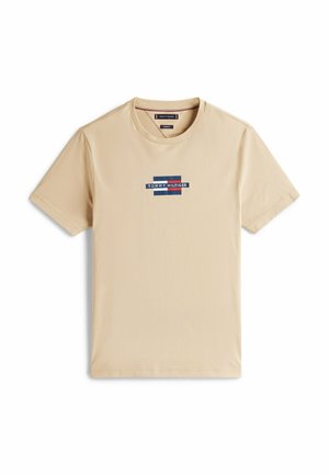 T-shirt di cotone beige con maniche corte, caratterizzato da una toppa logo blu e rossa centrata sul petto. Collo a giro, texture liscia.