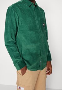 Zelená kordurová košile s límečkem na knoflíky, přední kapsou, texturovanými vertikálními pruhy a značkovou etiketu. Kombinováno s béžovými kalhotami.