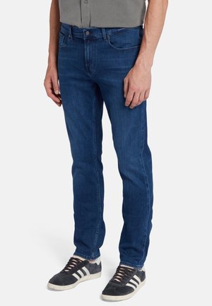 Jean en denim bleu, coupe slim, avec un design classique à cinq poches, présentant un léger effet délavé et des coutures contrastantes. Portés avec des baskets noires.