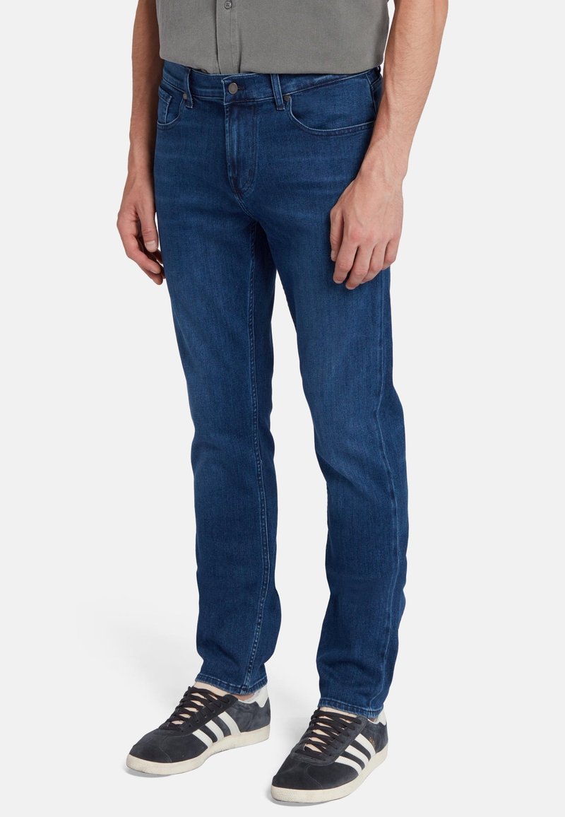 Jeans in denim blu, slim fit, con un classico design a cinque tasche, caratterizzati da una leggera sfumatura e cuciture in contrasto. Indossati con sneakers nere.