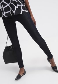 Svarta skinny jeans med en slät textur. I kombination med en mönstrad topp, spetsiga platta skor och en liten svart bucket-väska.