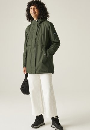 Kobieta o kręconych, ciemnych włosach, ubrana w oliwkowozieloną kurtkę z kapturem, białe spodnie, czarne buty i trzymająca małą czarną torbę.