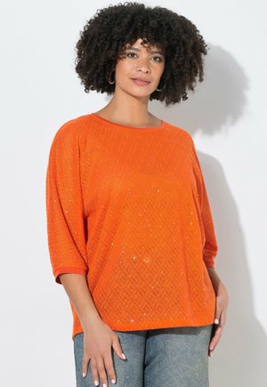 Kvinde med krøllet hår iført en lyseorange mønstret strikbluse med trekvartærmer og lyseblå jeans, stående mod en hvid væg.