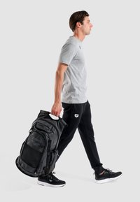 Mochila deportiva de malla y tela negra con múltiples compartimentos, llevada por una persona que viste una camiseta gris y pantalones deportivos negros.