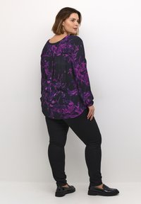 Langarm blouse in zwart met paarse bloemenpatroon, losse fit, ronde hals en geplooide achterkant. Gecombineerd met zwarte skinny jeans en glanzende instappers.