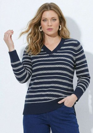 Kvinde med bølget blondt hår iført store hoop-øreringe, en navyblå og hvidstribet V-hals sweater og blå bukser med den ene hånd i lommen.