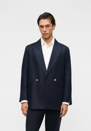Mand iført oversized marineblå dobbeltbrystet blazer med guldknapper over hvid skjorte, stående mod en ensfarvet lys baggrund.